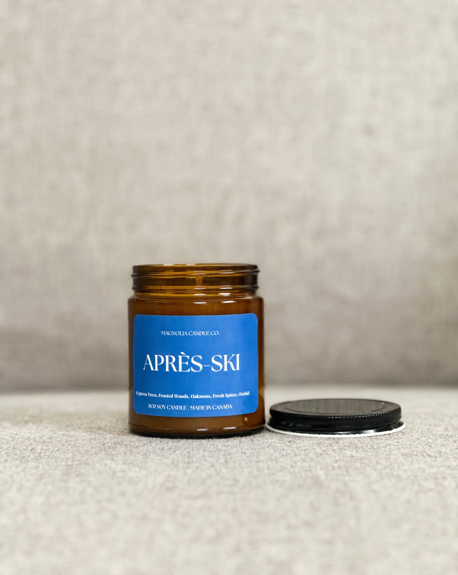 A lit amber glass jar candle with a blue label reading 'APRÈS SKI' sits on a rustic wooden table in a cozy indoor winter setting. A fireplace, snowy window view, gloves, and a mug are visible in the background.