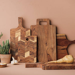 A set of wooden cutting boards and serving boards in various shapes and wood patterns, styled with kitchen props like potted cacti, garlic, a parsnip, and a small bowl against a warm terracotta background.