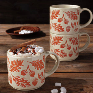Three ceramic mugs with an orange and beige autumn leaf and acorn pattern, shown on a rustic wooden table with hot chocolate, marshmallows, and a cinnamon stick.