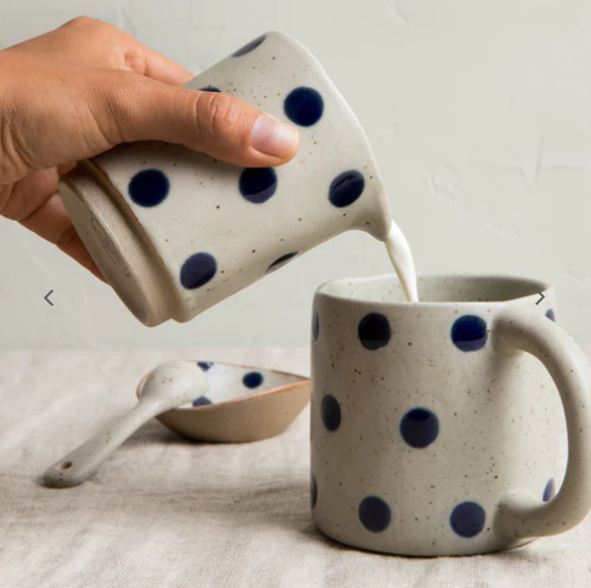 Person pouring milk from a polka dot ceramic pitcher into a matching mug on a light surface.
