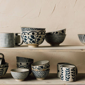 Collection of ceramic bowls and mugs with black patterns on a wooden shelf against a beige wall.