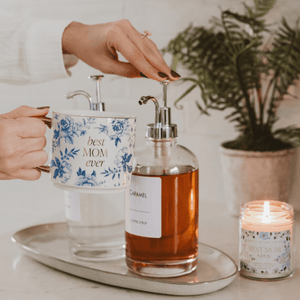 Caramel syrup bottle, floral mug with 'best mom ever' text, and lit candle on a tray with a plant in the background.