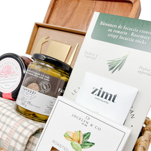 Assorted gourmet items including a bottle of olive oil, a jar of honey, and a box of chocolates on a wooden tray.