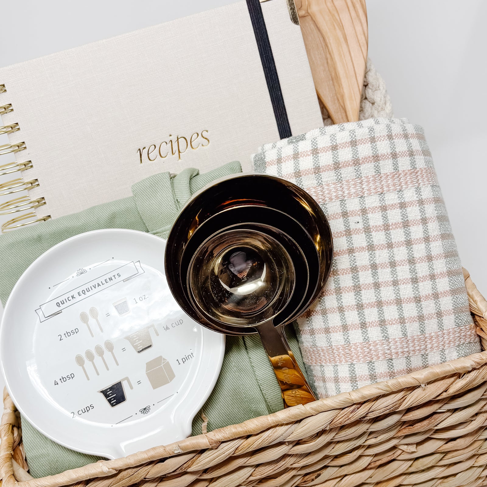 Baking set with a basket containing a cookbook, measuring cups, spoon, and towel on a light gray background.