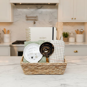 Baking set with a basket containing a cookbook, measuring cups, spoon, and towel on a light gray background.