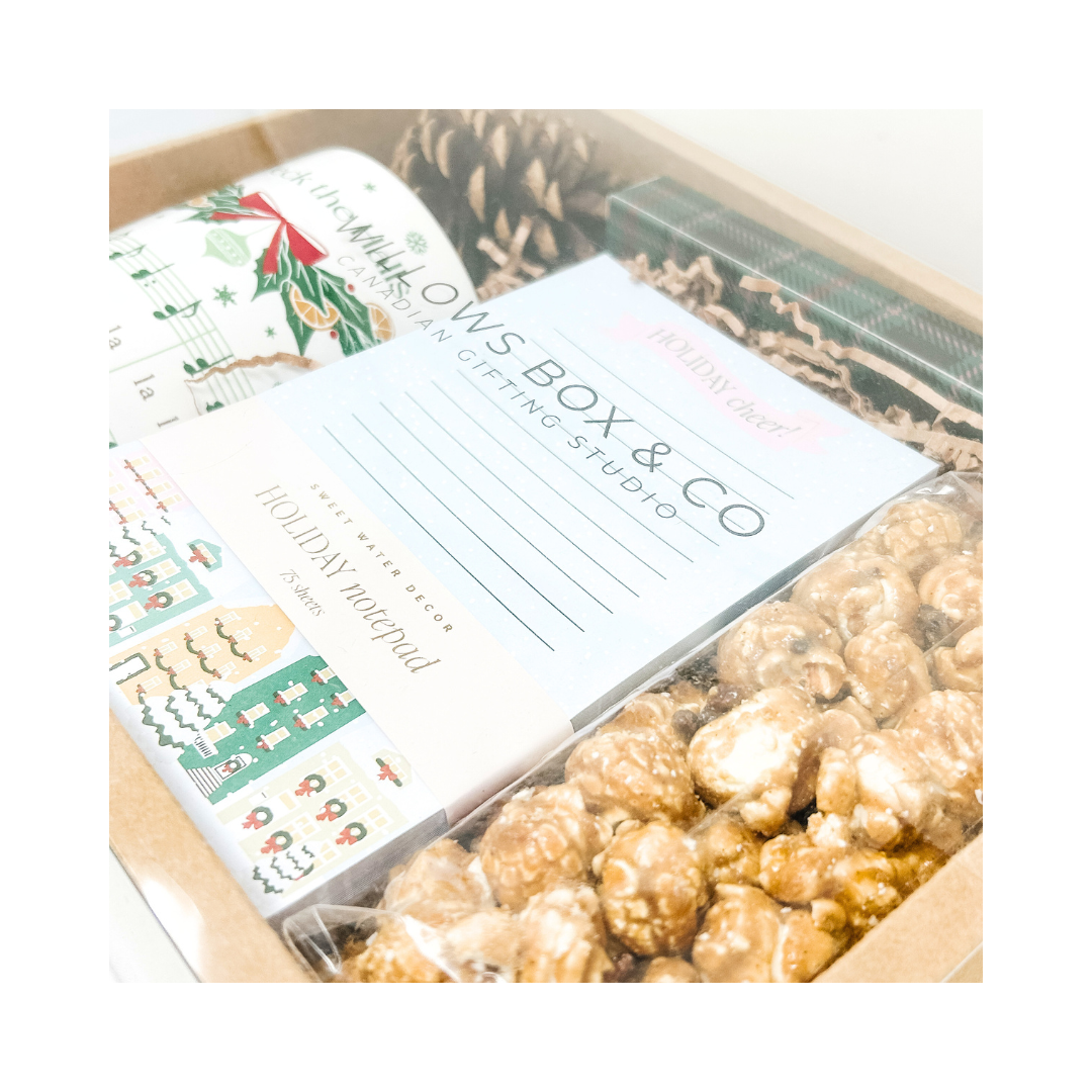 Gift set with holiday mug, holiday pen set, holiday note pad, pine cone, and caramel and chocolate popcorn in a kraft box, featuring 'Willows Box & Co.' branding.
