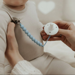 Baby sitting on a couch with a person holding a white pacifier and blue teething ring.