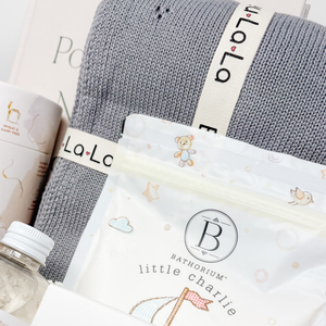 Gray knit blanket with 'La La' label, white bag with 'Bathorium little charlie' label, and water bottle on a magazine background.