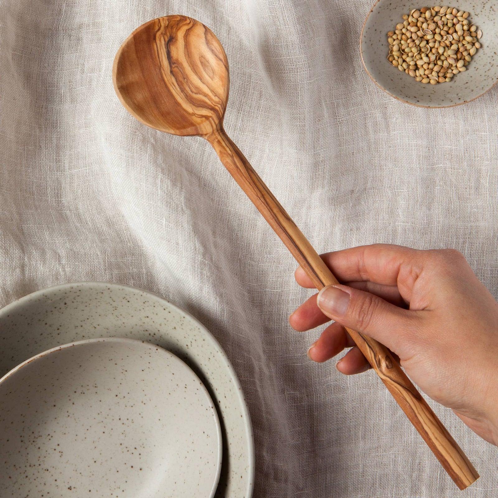 Hand holding a wooden spoon over ceramic bowls and a small dish of seeds on a textured surface.