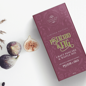 Purple box of 'Pistachio & Fig' craft pancake and waffle mix with figs and dried herbs on a white background