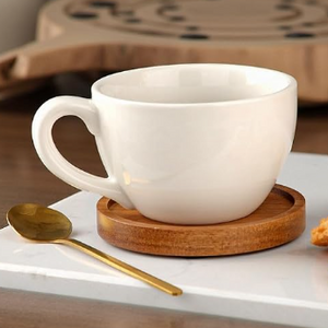 A white ceramic coffee cup with a rounded shape sits on a round wooden coaster, placed on a white marble serving board with a gold spoon and cookies nearby.