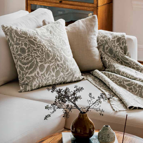 A styled living room scene with a cream sofa adorned with two patterned throw pillows, a matching patterned throw blanket draped over the seat, and a small round wooden coffee table in front holding a vase with dried stems and small decor objects.