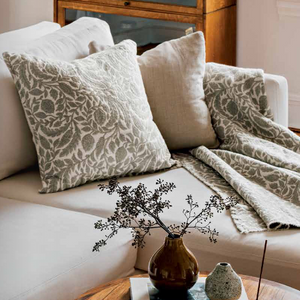 A styled living room scene with a cream sofa adorned with two patterned throw pillows, a matching patterned throw blanket draped over the seat, and a small round wooden coffee table in front holding a vase with dried stems and small decor objects.