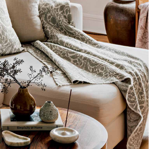 A styled living room scene with a cream sofa adorned with two patterned throw pillows, a matching patterned throw blanket draped over the seat, and a small round wooden coffee table in front holding a vase with dried stems and small decor objects.