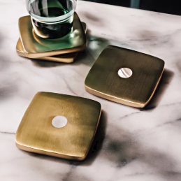 Three square brass coasters with circular cutouts, arranged on a white marble surface with a glass tumbler resting on one coaster.
