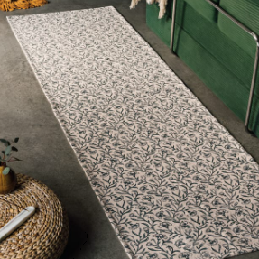 A long runner rug with an all-over botanical leaf pattern in dark gray on a warm beige background, styled in a modern living room next to a green sofa.