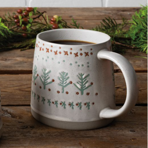 A ceramic mug filled with a hot beverage sits on a rustic wooden table, decorated with a light gray and white knit-style holiday pattern of green pine trees, red berries, and small stars. A second mug is partially visible on the left, and evergreen branches with red berries decorate the background against a white brick wall.