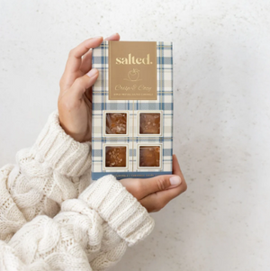 A person in a cream cable-knit sweater holds a box of 'salted.' brand Caramel & Cream gourmet caramel candies against a light, textured background.