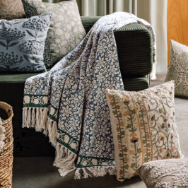 A patterned throw blanket with fringe is draped over a dark sofa in a living room setting, surrounded by matching decorative pillows.