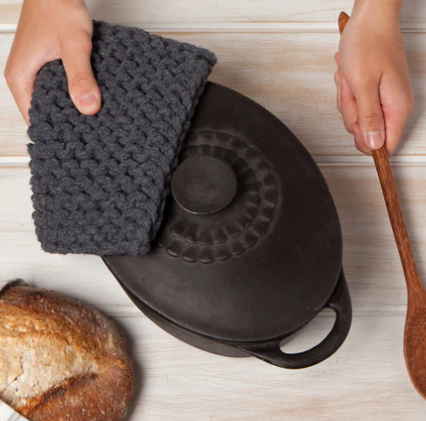 Person holding a dark gray textured cloth over a black cast iron skillet on a light wooden surface with a wooden spoon and bread.