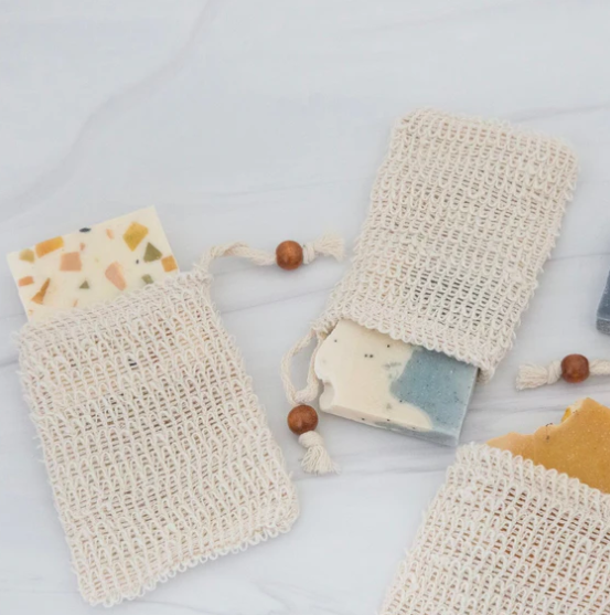 A beige sisal soap saver bag with a drawstring, featuring a label that reads 'SUNTREE SOAPS Exfoliating Soap Saver' and is placed on a wooden surface with a dark background.