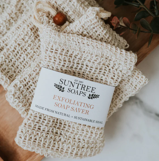 A beige sisal soap saver bag with a drawstring, featuring a label that reads 'SUNTREE SOAPS Exfoliating Soap Saver' and is placed on a wooden surface with a dark background.