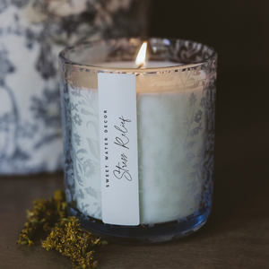 Candle with a label on a wooden surface, surrounded by flowers.