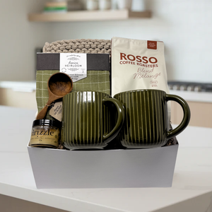 Gift basket with green mugs, coffee, and a wooden spoon on a white surface.