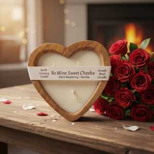 Heart-shaped candle labeled 'Be Mine Sweet Cheeks' next to a bouquet of red roses on a wooden surface.
