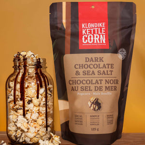Klondike Kettle Corn dark chocolate sea salt popcorn package with a jar of popcorn on a wooden surface.