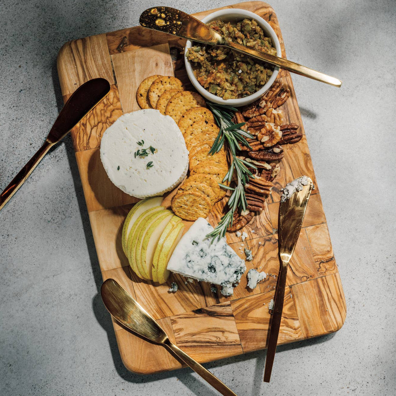 Wooden cutting board with assorted cheeses, crackers, and accompaniments on a gray surface