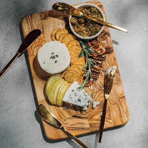 Wooden cutting board with assorted cheeses, crackers, and accompaniments on a gray surface