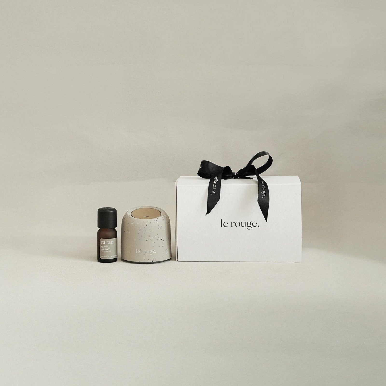A gift set from le rouge. featuring a white box tied with a black ribbon, a small beige ceramic diffuser, and a dark amber essential oil bottle, set against a neutral warm beige background.