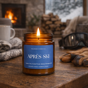 A lit amber glass jar candle with a blue label reading 'APRÈS SKI' sits on a rustic wooden table in a cozy indoor winter setting. A fireplace, snowy window view, gloves, and a mug are visible in the background.